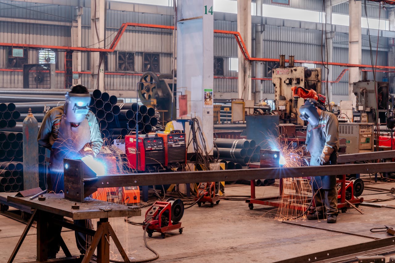 services-03 Two welders wearing protective gear working in a busy industrial factory workshop.